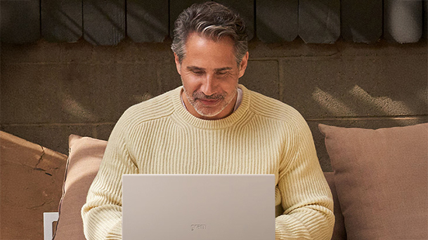 The image shows a man in a yellow sweater sitting on a couch working on a laptop with a copy of "Top businesses have already deployed new Windows 11 Pro PCs."