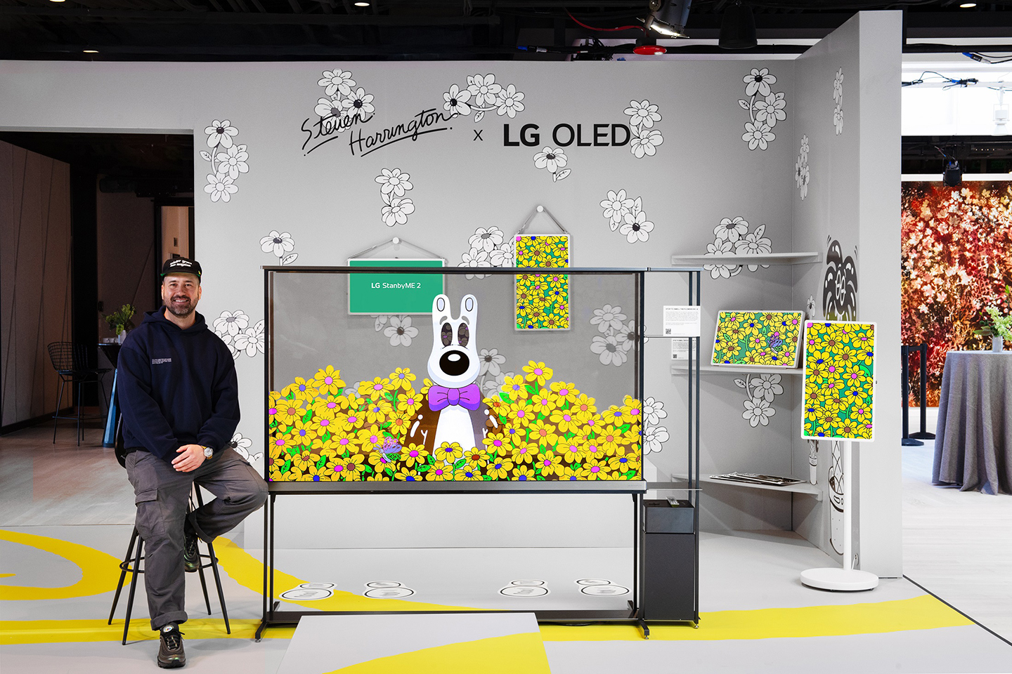 Artist Steven Harrington is sitting on a stool beside a transparent LG OLED TV that displays his signature artwork in a themed exhibition space.