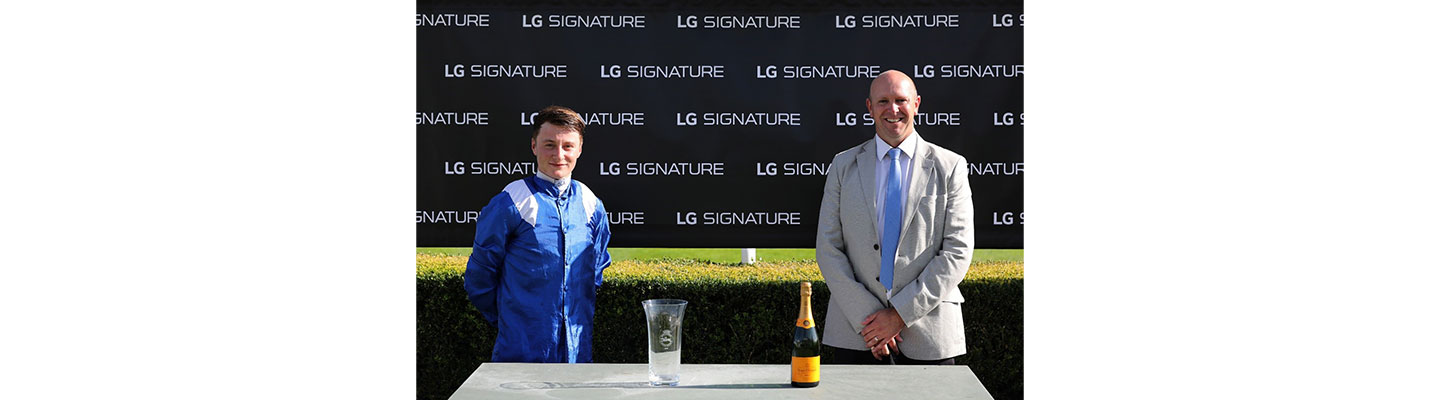 A winning jockey from the Qatar Goodwood Festival is presented with a glass trophy and a bottle of champagne in front of an LG SIGNATURE-branded sign