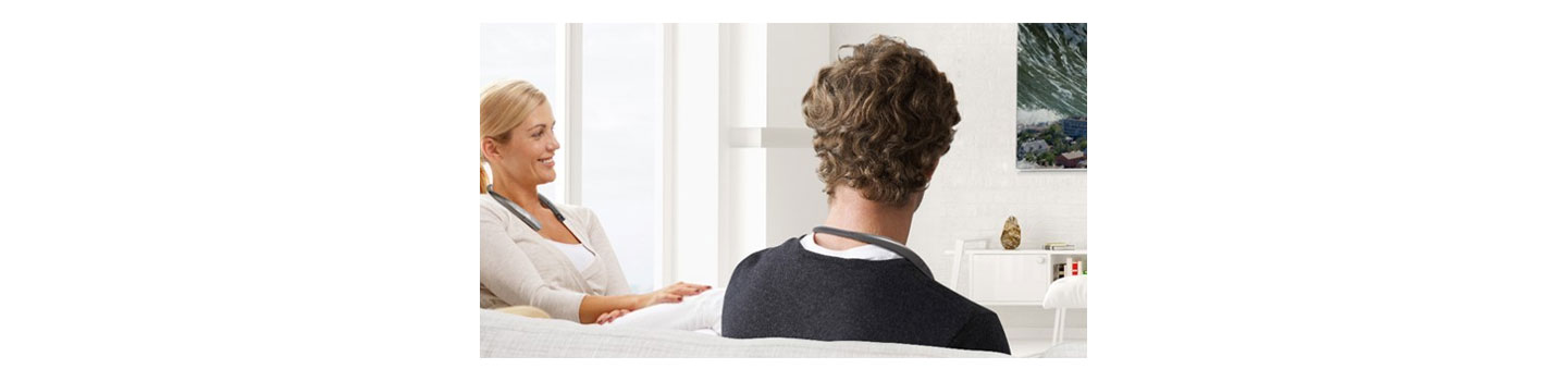 A man and a woman sit on a couch and talk to each other while wearing the LG TONE Studio Bluetooth wearable speaker.