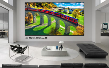 An ultra-large LG Micro RGB evo TV is mounted to the wall in a modern living room, with a vivid red train crossing a stone bridge displayed on the screen.