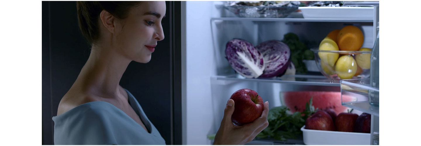 A woman takes an apple from her fruit and vegetable-filled LG refrigerator that keeps ingredients fresher for longer with HygieneFRESH+™ and LINEARCooling™ technology