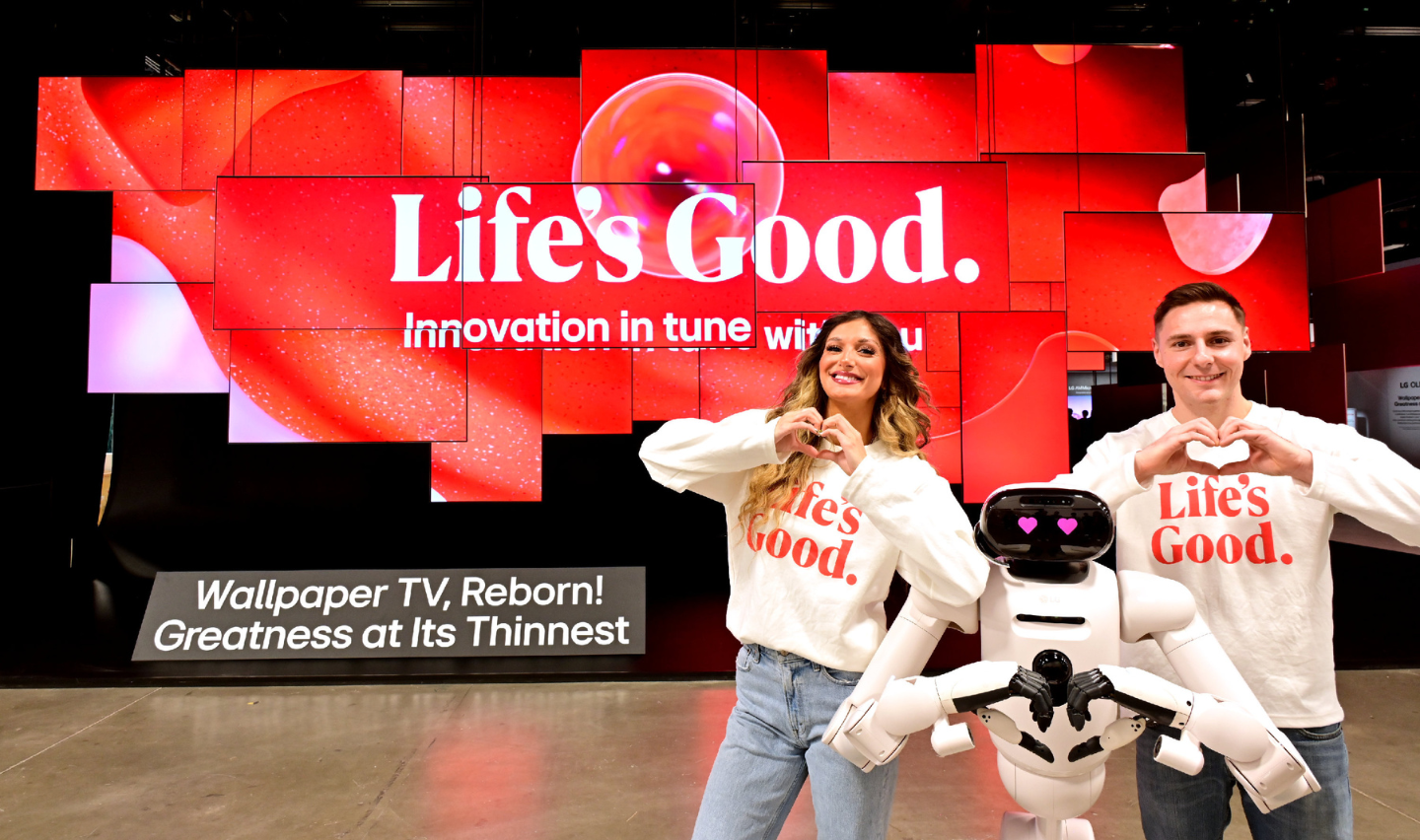 A man and woman pose with the LG CLOiD home robot, all making a heart shape with their hands in front of the In Tune Monument at the booth entrance.