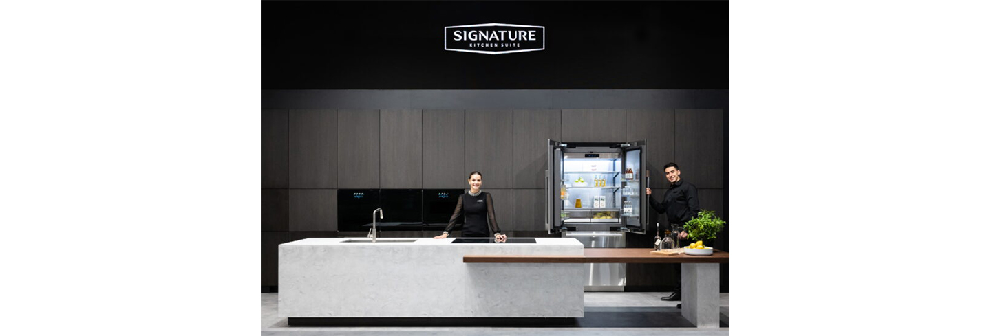 A woman is standing behind the kitchen island and a man is holding the Signature Kitchen Suite built-in 36-inch French Door refrigerator door at LG booth