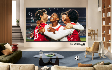 Four soccer players are celebrating a goal on the screen of a wall-mounted LG QNED evo Mini LED TV in a bright living room.