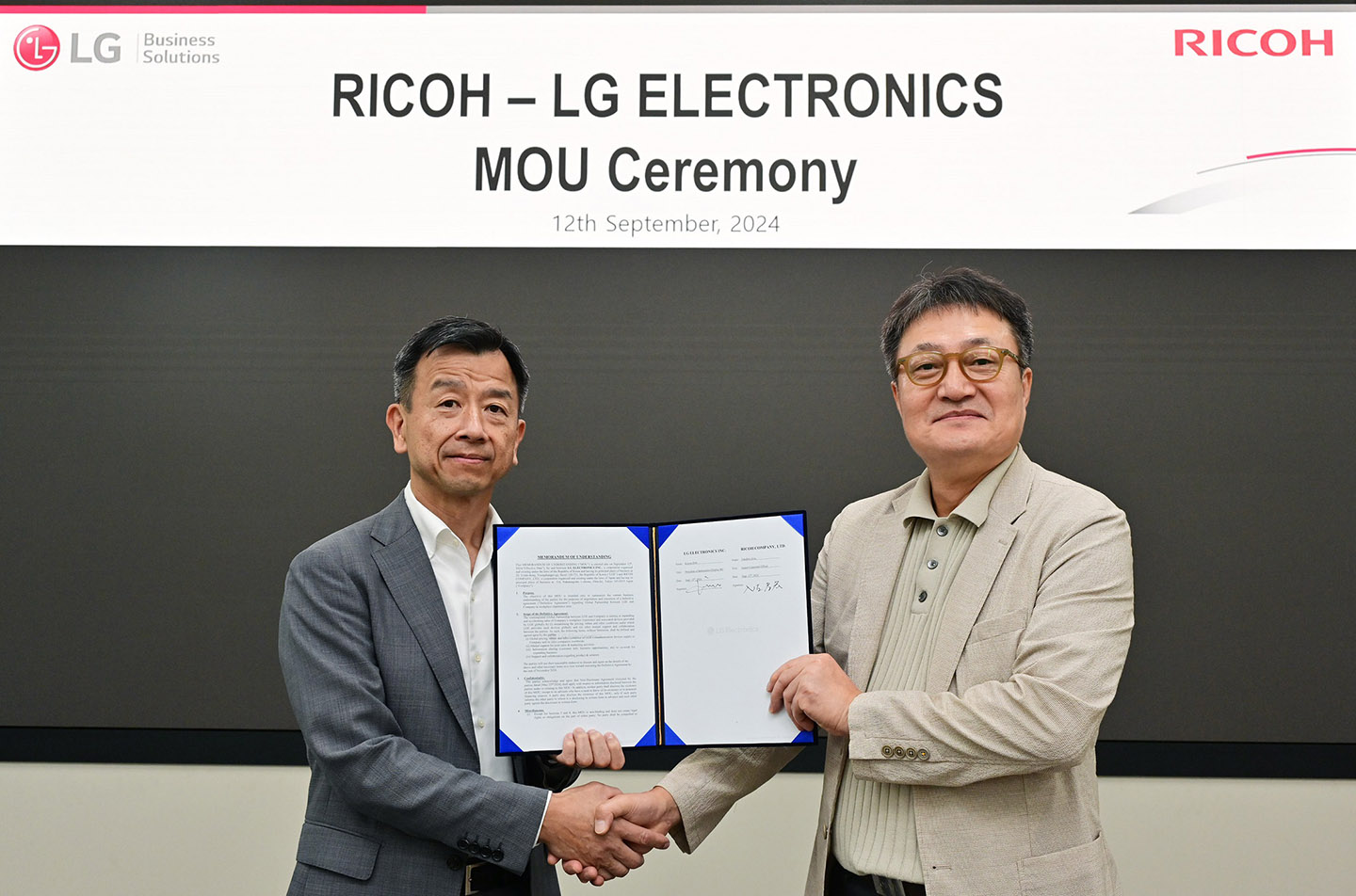 Representatives from LG (left) and Ricoh (right) shake hands while holding a signed MOU