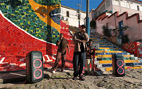 A group of three street performers in Brazil deliver a vibrant performance in front of a colorful mosaic mural and staircase, accompanied by two LG XBOOM speakers