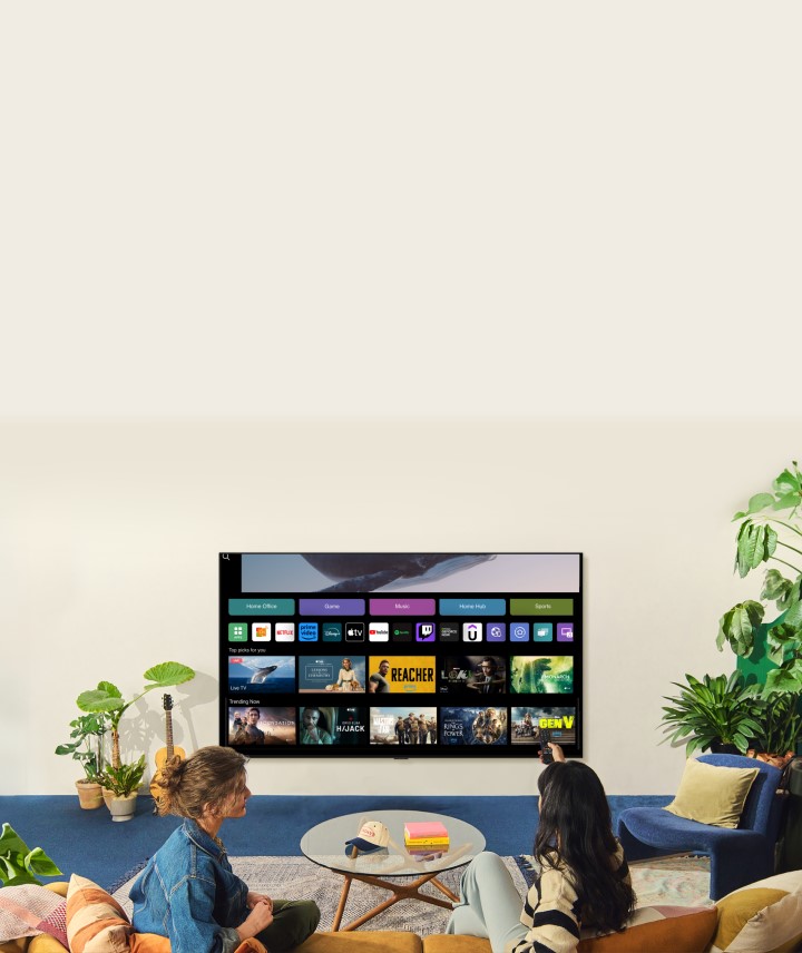 A whale floating above the ocean with a woman in the ocean are shown. A home screen appears from below. As the scene transitions, it shows the image in a large LG TV on the wall. Two women sit in a cozy and neutral living room filled with plants, and a guitar. One woman points the remote at the TV, which shows a range of apps and recommended TV shows on the home screen.