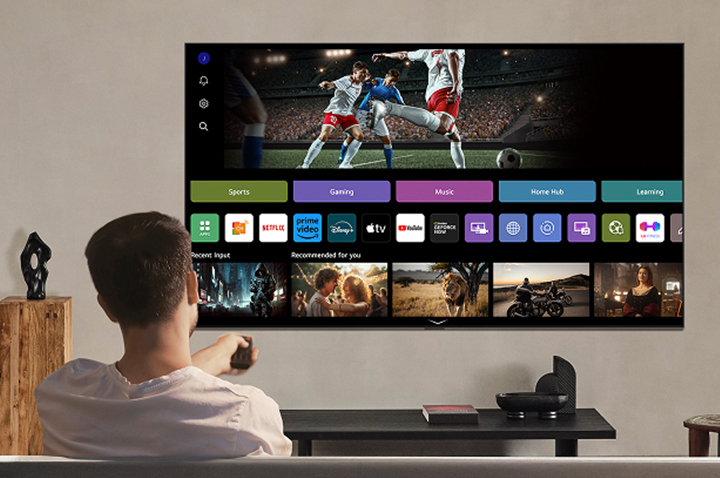 Person on the couch holding out a remote control. LG AI TV on the wall with LG webOS on the screen.