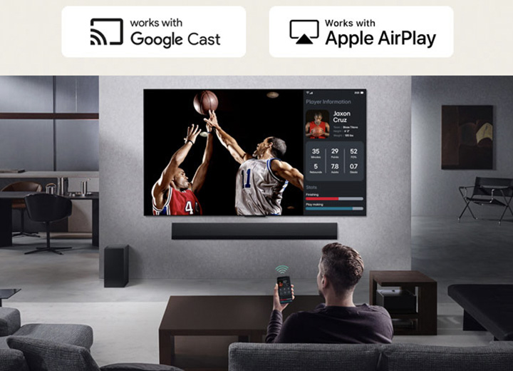 Person in their living room holding their phone. On the phone is a transmitting icon showing that the phone screen is being mirrored on the TV. On the TV is a basketball game to the side is the mirrored screen, showing player statistics.