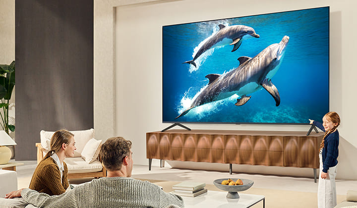 A girl and a dog are seated in front of a LG QNED TV mounted on a wall depicting three elephants walking outwards above a LG Soundbar. 