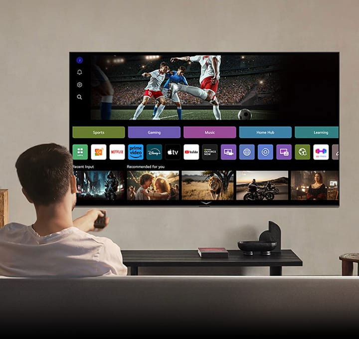 Person on the couch holding out a remote control. LG AI TV on the wall with LG webOS on the screen.