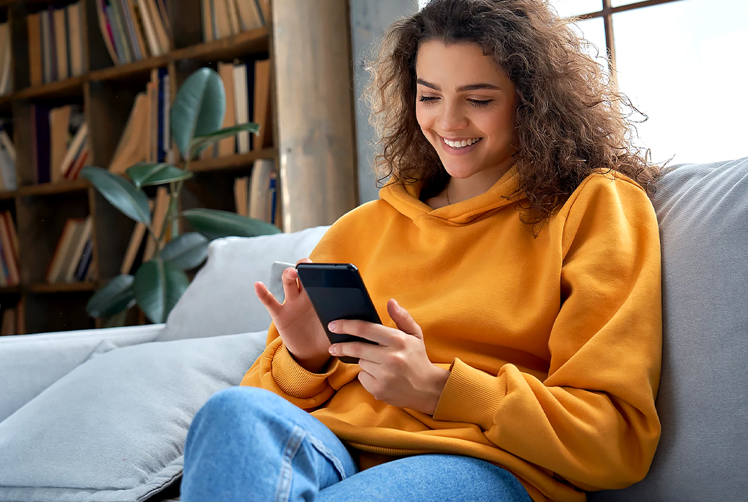 Woman smiling at her phone on a sofa illustrates Smart Diagnosis support for checking LG laundry product status through an app.