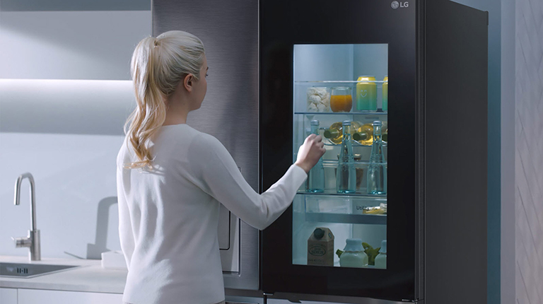 A woman in a white jumper opens an LG refrigerator door in a sleek kitchen, revealing neatly arranged food and drinks.