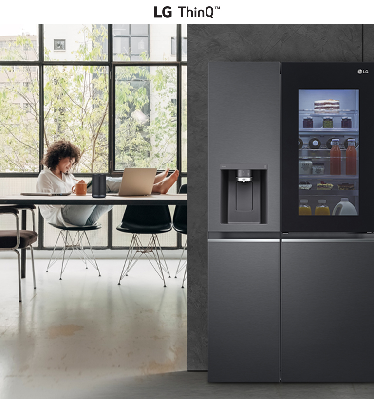 Modern kitchen with an LG ThinQ refrigerator on the right and a person using a laptop by a large window on the left.