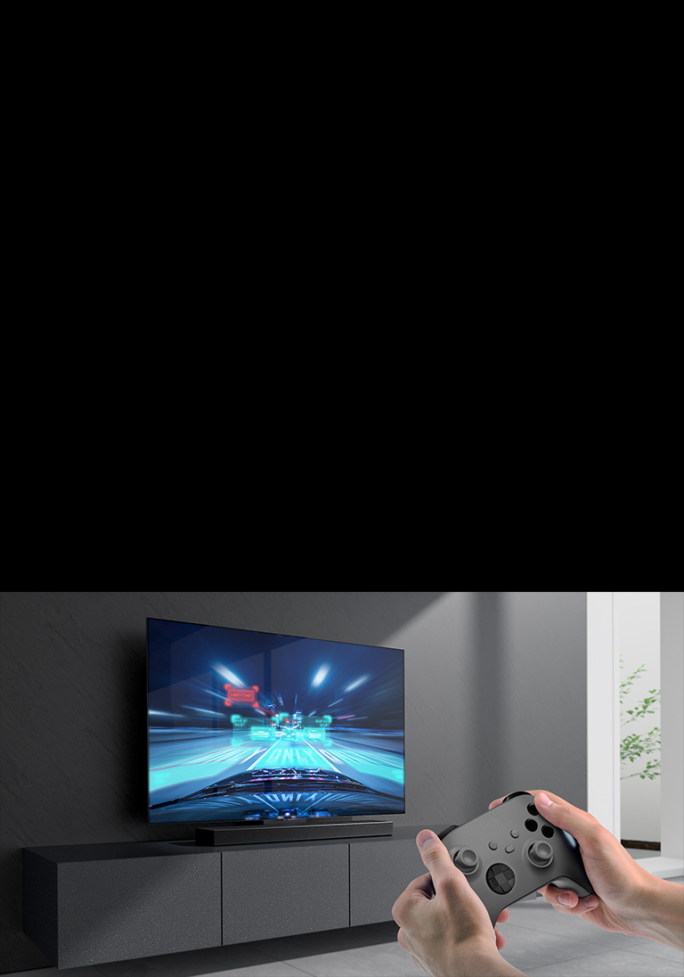 The LG Soundbar is placed on the cabinet and racing game scene is shown on the LG TV coneected to the Soundbar . A game console is on the bottom right side of the picture hold by two hands.