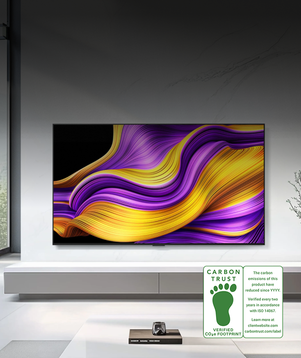 An image of an LG OLED TV product. An LG OLED TV is wall-mounted on a white wall, displaying an abstract graphic with purple and yellow wave patterns on screen. In the lower right, a CARBON TRUST VERIFIED CO₂e FOOTPRINT certification mark is shown along with the text: 'The carbon emissions of this product have reduced since YYYY. Verified every two years in accordance with ISO 14067. Learn more at carbontrust.com/label.'
