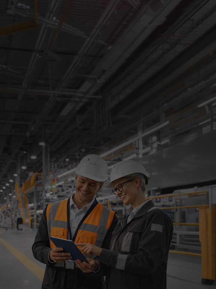 A spacious, brightly lit manufacturing plant interior equipped with industrial machinery. On the right, two workers wearing white hard hats, safety glasses, and high-visibility workwear are looking together at a tablet device.