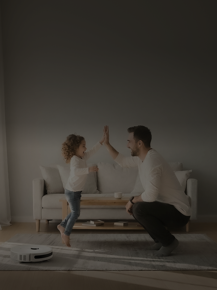 A smiling man and a child standing on a coffee table high-fiving in a bright living room, with a robot vacuum on the floor