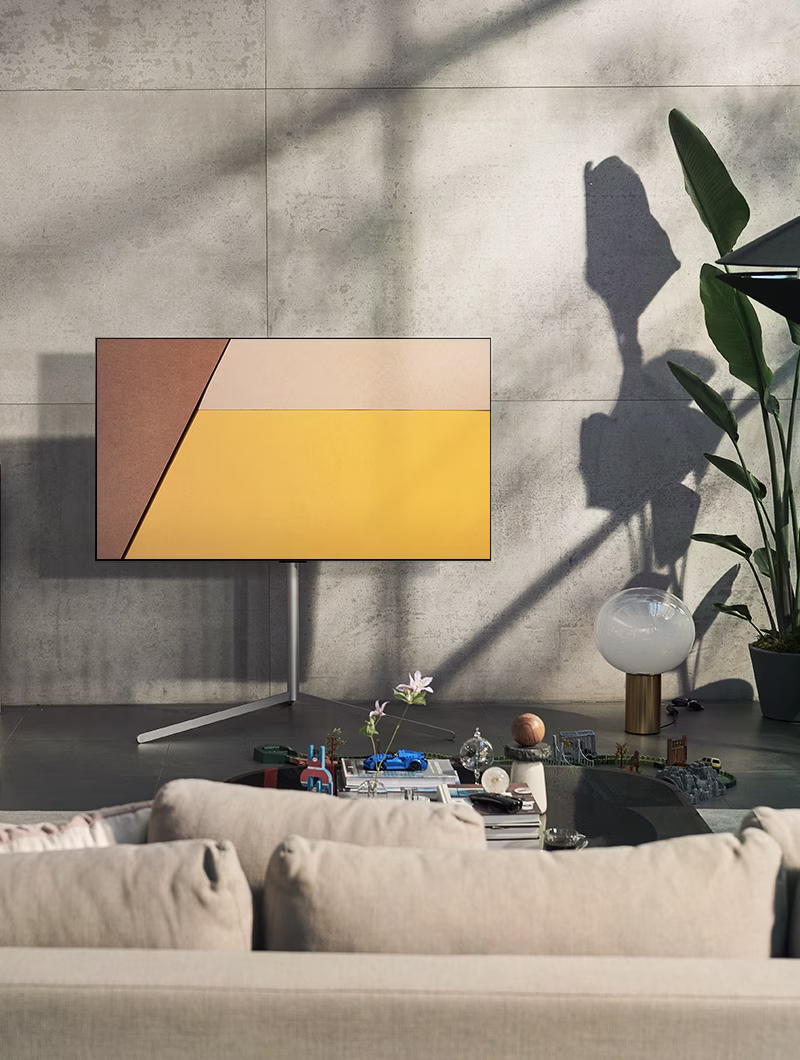An LG OLED television is in a modern gray living space against a window showing a swimming pool. An LG OLED television is in a neutral living space with exposed bricks, wooden details, and greenery.