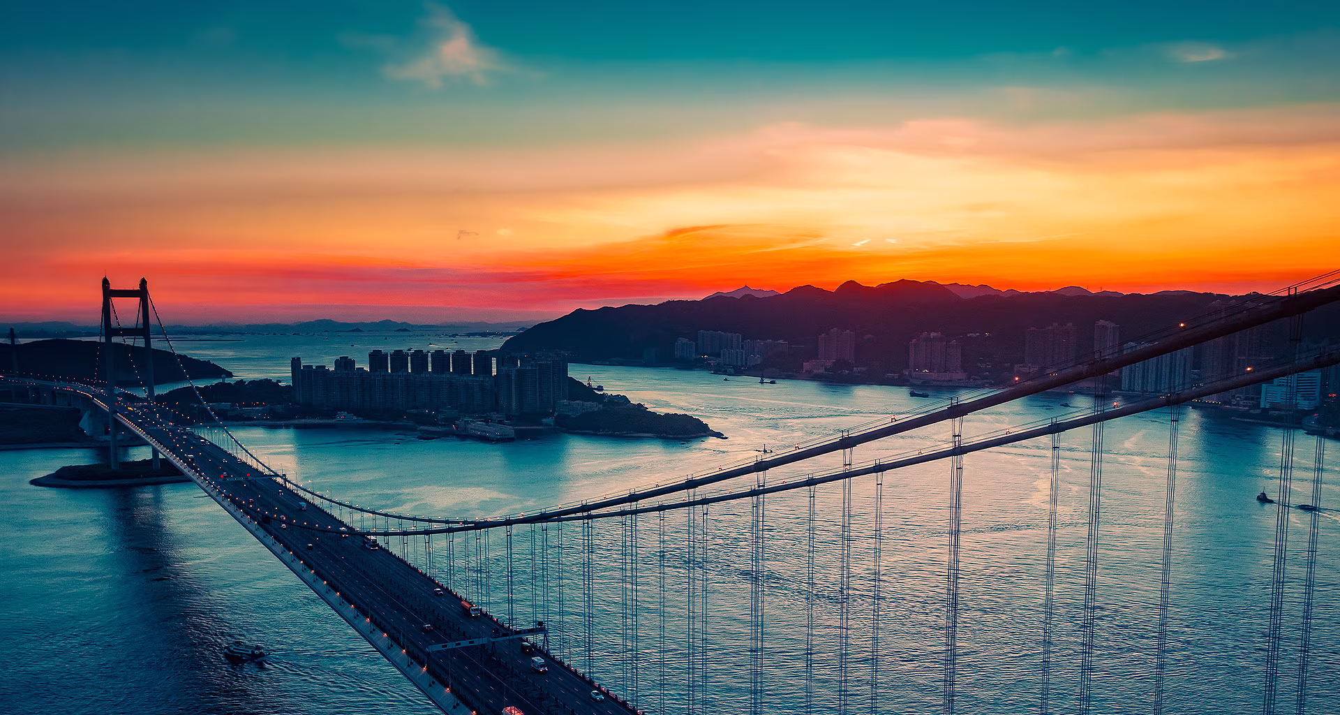 An ariel view of a coastal city with traffic driving over a bridge as the sun sets appears across the full screen. It is cropped and shown on an LG OLED television screen with the original colors unchanged.	  