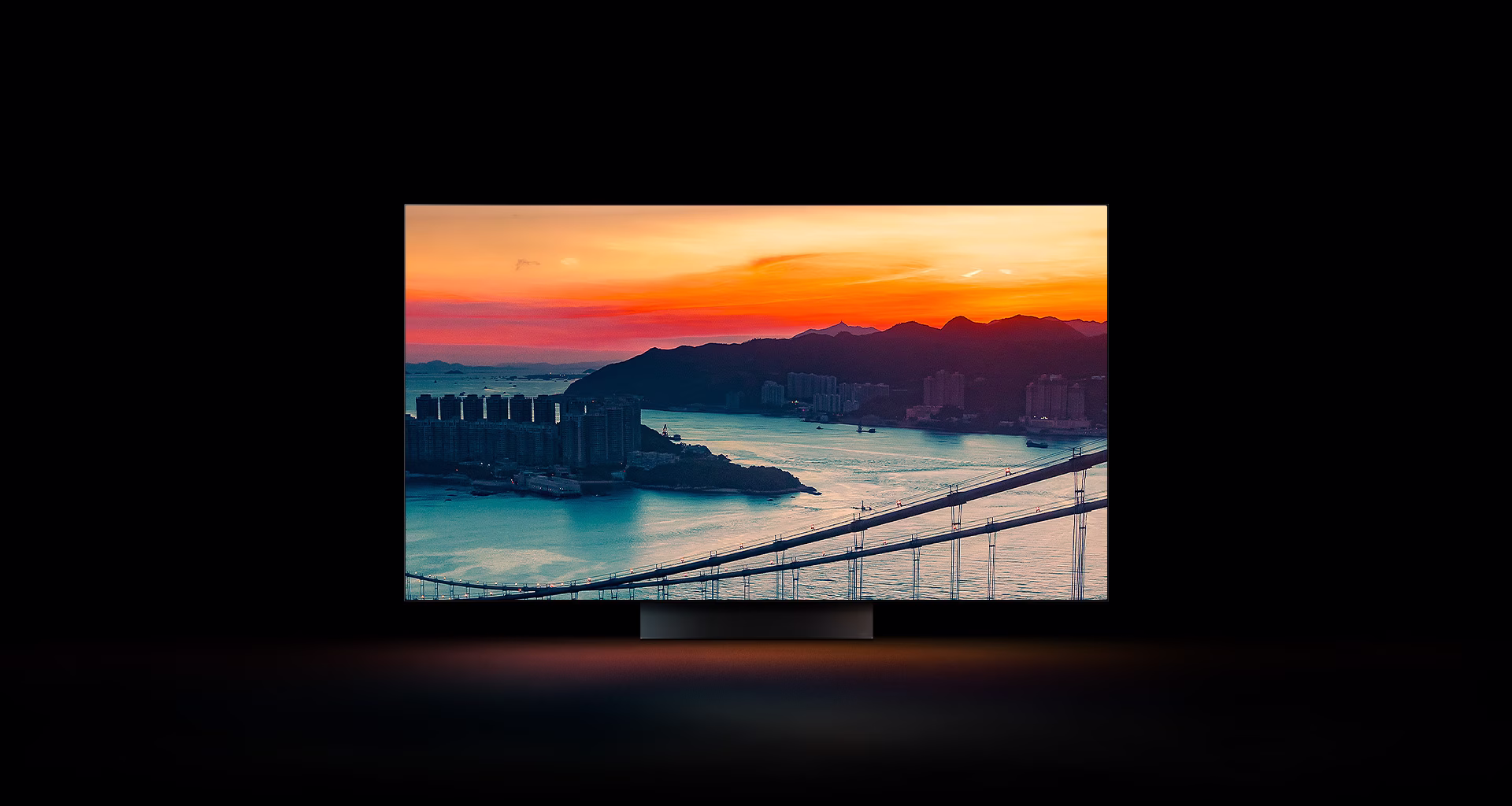An ariel view of a coastal city with traffic driving over a bridge as the sun sets appears across the full screen. It is cropped and shown on an LG OLED television screen with the original colors unchanged.	  